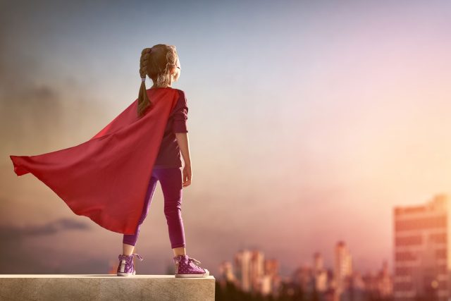 a little girl wearing a red cape standing on a ledge.